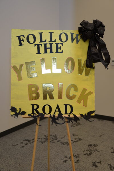 A colorful sign reading 'Follow the Yellow Brick Road' with decorative elements and a black bow, perfect for themed events.